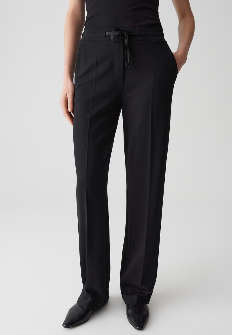 Pantaloni neri su misura con una vita regolabile, frontale piatto, tasche laterali e una texture liscia. Indossati con scarpe nere a punta.