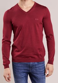 Röd långärmad v-neck tröja i finstickad kvalitet. Har en diskret logo på bröstet samt ribbade muddar och nederkant. Matchas med blå jeans.