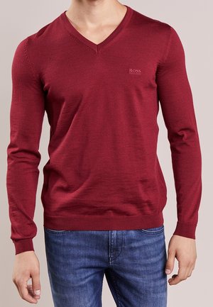 Roter langärmliger V-Ausschnitt-Pullover aus feinem Strickstoff. Mit einem subtilen Logo auf der Brust sowie gerippten Bündchen und Saum. Kombiniert mit blauen Jeans.