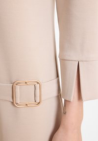 Beige Stoffärmel mit einem Schlitz am Bündchen und passendem Gürtel mit quadratischer, matter goldfarbener Schnalle an einem hellen Arm, der ein kleines Tattoo zeigt.