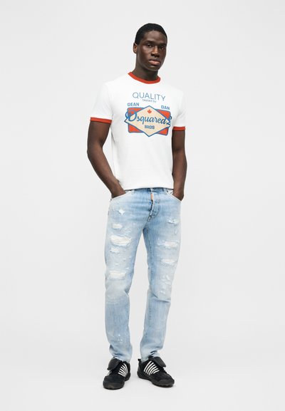 Homme debout les mains dans les poches, portant un t-shirt blanc à motif, un jean clair déchiré et des baskets noires avec des rayures blanches.