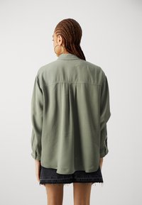 Olive green, long-sleeve shirt with a relaxed fit, featuring a back pleat. Paired with black denim shorts, frayed at the edges.