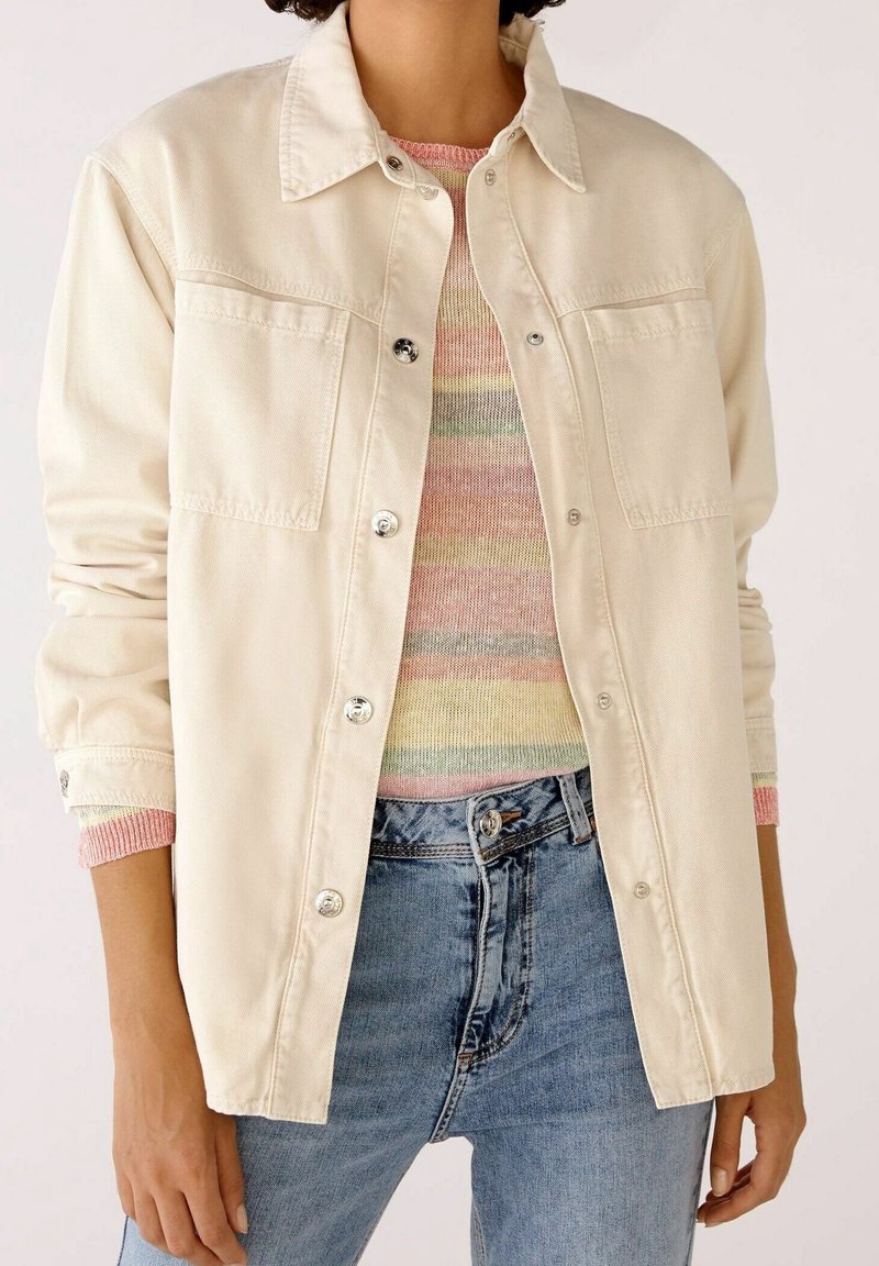 Giacca di jeans beige con chiusura a bottoni, due tasche sul petto e maniche arrotolate su un maglione a righe multicolore. Visibili jeans azzurri.