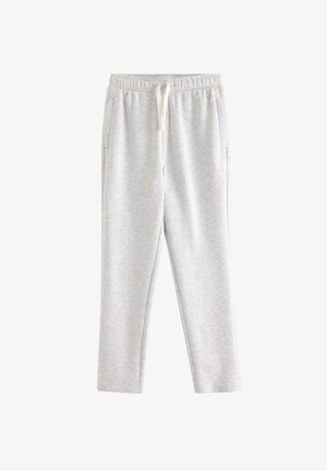 Jongen met rood haar die een witte kabelgebreide trui, een marineblauw shirt, lichtgrijze sweatpants en crèmekleurige sneakers draagt, staand met één hand achter zijn hoofd.