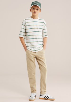 Dječak s zelenom kapom, bijelom majicom na pruge, bež hlačama i bijelim tenisicama s crnim prugama stoji s rukama u džepovima.