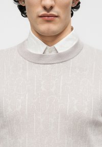 Κοντινό πλάνο ενός άνδρα που φοράει ένα ανοιχτό μπεζ πουλόβερ με σχέδιο πάνω από ένα λευκό πουκάμισο με κουμπιά, με εμφανή σκουλαρίκια με καρφίτσες.