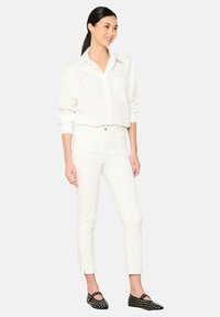 Camisa blanca de botones con un solo bolsillo en el pecho, combinada con jeans ajustados blancos. Zapatos planos negros con tachuelas plateadas. Tela suave, ajuste casual.