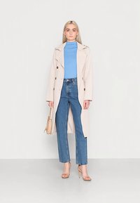 Licht beige trenchcoat met dubbelgeknoopte knopen, gedragen over een lichtblauwe coltrui en hoog uitgesneden blauwe spijkerbroek. Doorzichtige sandalen met blokhak.