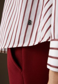 Camisa a rayas blanca y burdeos con un pequeño logo negro, combinada con pantalones burdeos ajustados que tienen un detalle de bolsillo lateral.