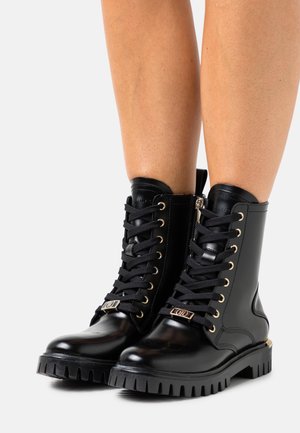 Černé kožené kotníkové boots na šněrování s akcenty ze zlaceného kovu, robustní podrážkou a elegantním designem. Obsahují zip a texturované detaily.