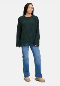 Dunkelgrüner strukturierter Pullover mit lockerer Passform, fransenbesäumtem Saum und langen Ärmeln, kombiniert mit hellblauen Jeans und beigen Schuhen.