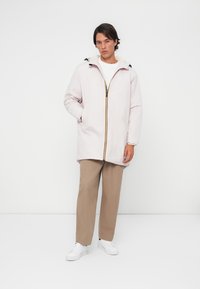 Veste rembourrée rose clair avec capuche, fermeture éclair complète et finitions jaunes contrastantes. Le modèle porte un pantalon beige et des baskets blanches.