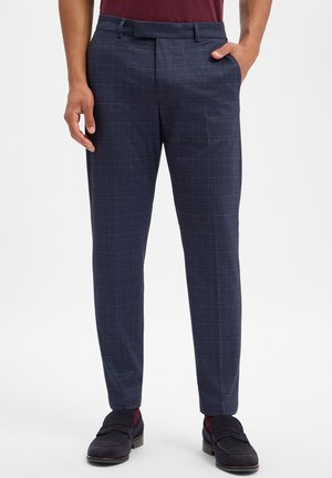 Man draagt donkerblauwe geruite slim-fit broek met zwarte loafers en bordeauxrode sokken, staat met één hand in zak.