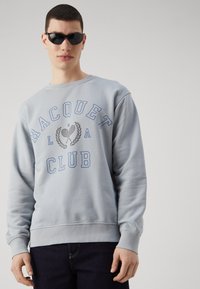 Sudadera azul claro con el texto "RACQUET CLUB" en azul, con puños y dobladillo de canalé. Llevada con gafas de sol negras en forma ovalada.