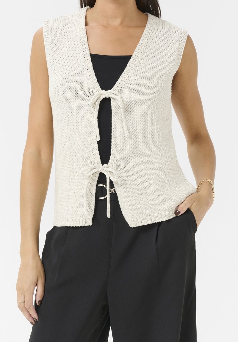Kvinde iført ærmeløs beige strikket vest bundet med to sløjfer over en sort top, parret med sorte højtaljede bukser og et guldarmbånd.
