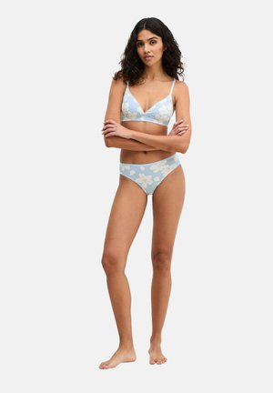 Woman standing barefoot with arms crossed, wearing light blue floral bra and matching underwear against plain white background.