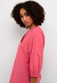 Blusa in cotone rosa con un motivo testurizzato, scollatura a V e maniche a tre quarti. Presenta un dettaglio arricciato ai polsini delle maniche.