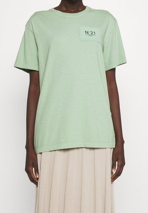 Osoba ubrana w jasnozieloną koszulkę z krótkim rękawem z małą kieszonką i napisem "N°21", połączoną z beżową plisowaną spódnicą.