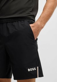 Melni sporta šorti, kas izgatavoti no viegla materiāla, ar elastīgu jostu, sānu kabatām un balto "BOSS" logotipu ar krāsainām svītrām.