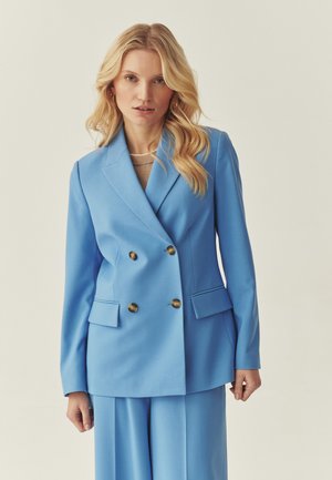 Blonde vrouw draagt een lichtblauwe dubbele rij blazer met gouden knopen en bijpassende wijde broekspijpen, staande tegen een effen achtergrond.