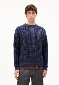 Navy blauwe gebreide trui met gestructureerde, verticale patronen, geribde kraag en manchetten, en een reguliere pasvorm. Model draagt een gelaagd outfit.