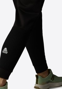 Melni sporta bikses ar teksturētu apakšējās kājas daļu, ar atstarojošu "Mountain Athletics" logo un kombinētas ar gaiši zaļiem sporta apaviem.