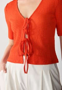 Haut rouge texturé avec un décolleté en V profond et des manches courtes, présentant une fermeture à nouer à l'avant et un motif en tricot ondulé, associé à un pantalon de couleur claire.