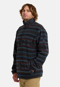Fleece trui in donkere tinten met blauwe en roestkleurige strepen, ritskraag en een borstzak. Heeft een zachte textuur en een ontspannen pasvorm.