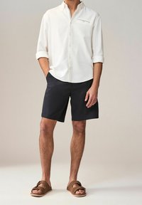 Chemise blanche à manches longues en tissu texturé, shorts noirs et sandales marron. La chemise présente un col boutonné et une rayure décorative.