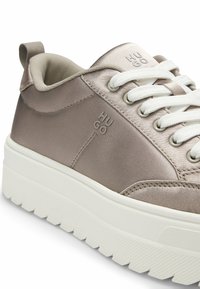 Light brown satin sneakers with white laces, featuring a thick white sole and embossed "HUGO" logo. Rounded toe and padded collar.