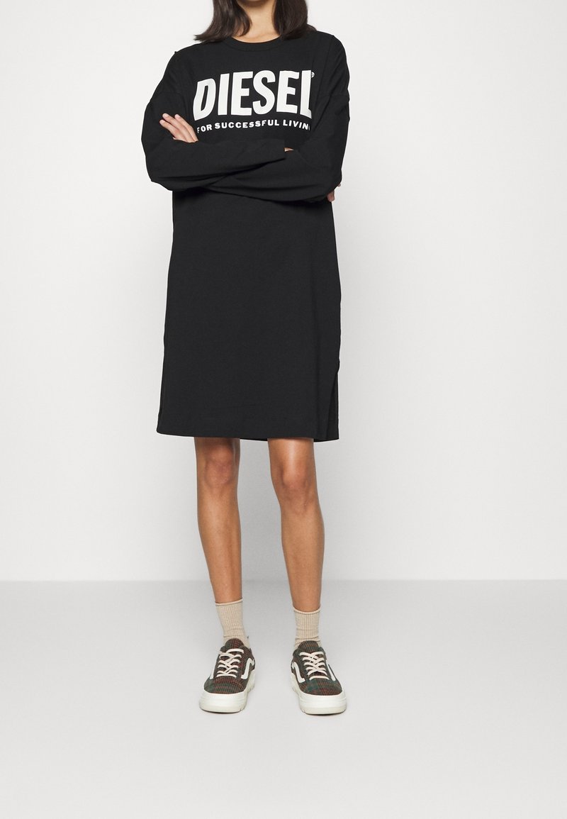 Personne portant une robe noire à manches longues et longueur genou avec le texte "DIESEL FOR SUCCESSFUL LIVING", des chaussettes beiges et des baskets vertes à motifs.