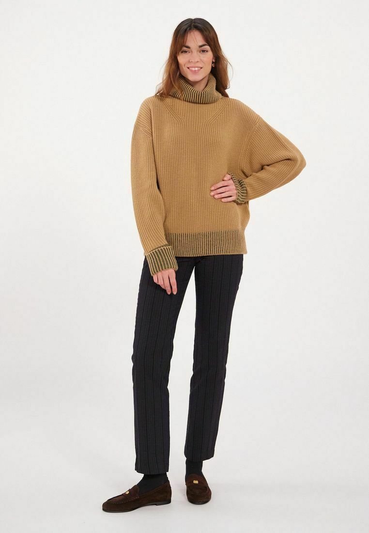Pull en beige à côtes avec un col montant, coupe oversize et poignets contrastants en vert foncé, associé à un pantalon rayé foncé et des mocassins marron.