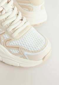 Scarpa da ginnastica beige e bianca con tomaia in mesh, cuciture tono su tono e suola strutturata. Presenta lacci piatti e dettagli di design sottili.
