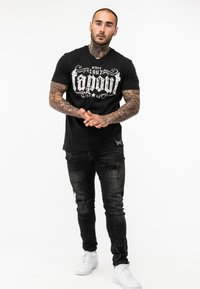 Fekete póló fehér "Tapout" logóval, elhasználódott fekete farmernadrággal és fehér sportcipővel párosítva, amely testéhez simuló sziluettet és rövid ujjakat mutat.