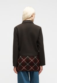 Άτομο με μακριά ξανθά μαλλιά φοράει σκούρο καφέ σακάκι πάνω από τοπ με κόκκινα και μαύρα διαμάντια και μπλε τζιν, φαίνεται από πίσω.