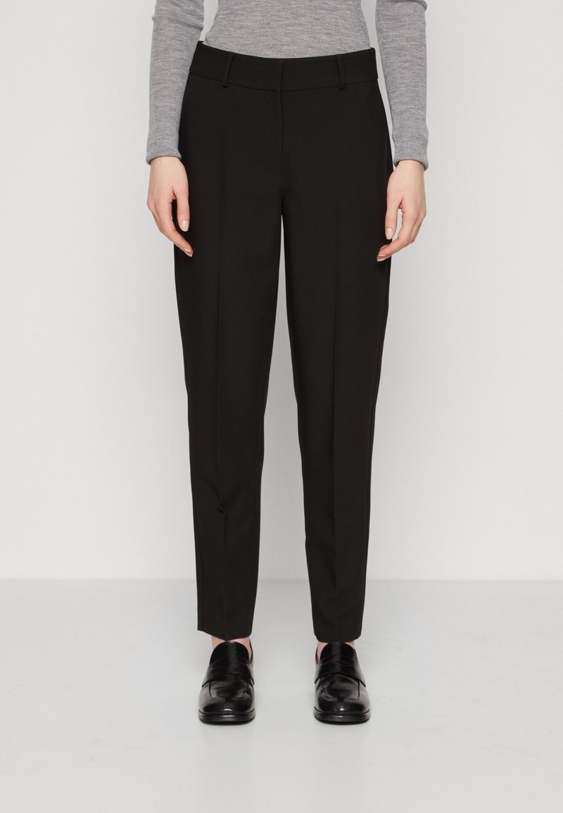 Pantalon noir sur mesure avec une texture lisse, coupe slim et taille standard. Associé à des chaussures noires, porté avec un haut gris à manches longues.