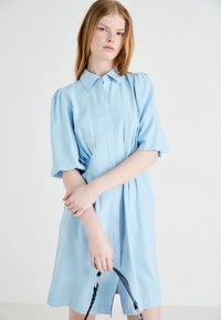 Robe bleu clair à manches courtes avec un col à boutons, des détails plissés et une coupe droite, portée par un modèle avec une bretelle tressée.
