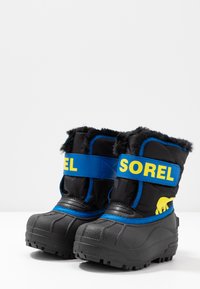 Schwarze Winterstiefel mit blauen Riemen, auf denen "SOREL" in Gelb steht. Mit einem Pelzbesatz an der Oberkante, strukturiertem Gummi-Sohle, wasserdichtem Design und modernem Aussehen.