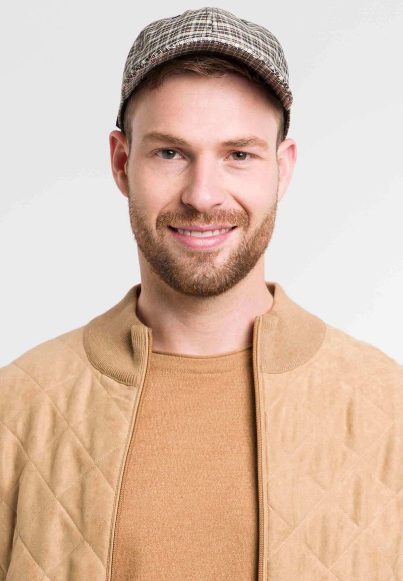 Beige gesteppte Jacke mit Stehkragen, über einem braunen Pullover getragen. Die Person trägt eine kariierte Mütze. Der Hintergrund ist neutral.