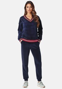 Marineblauer Velours-Trainingsanzug mit roten und weißen Streifenakzenten, bestehend aus einem V-Ausschnitt-Pullover und engen Jogginghosen mit elastischen Bündchen.