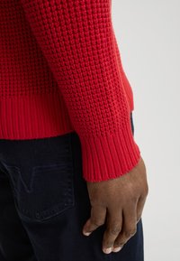 Rode gebreide trui met textuurpatroon en geribbelde manchetten, gecombineerd met donkerblauwe broek met contrasterende stiksels. Hand rust op het been.