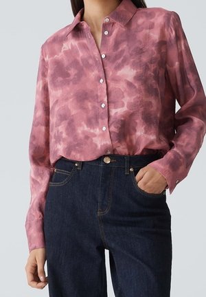 Frau trägt ein langärmliges rosa Batikhemden mit Knöpfen, das in hoch taillierte dunkelblaue Jeans gesteckt ist, mit einer Hand in der Tasche.