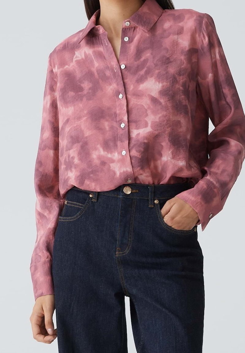 Femme portant une chemise rose tie-dye à manches longues boutonnée, rentrée dans un jean bleu foncé taille haute, avec une main dans une poche.
