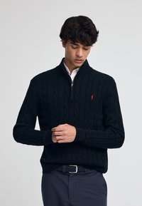 Pull noir en tricot torsadé avec un col zippé, arborant un petit logo rouge sur la poitrine et des poignets côtelés, associé à un pantalon foncé.