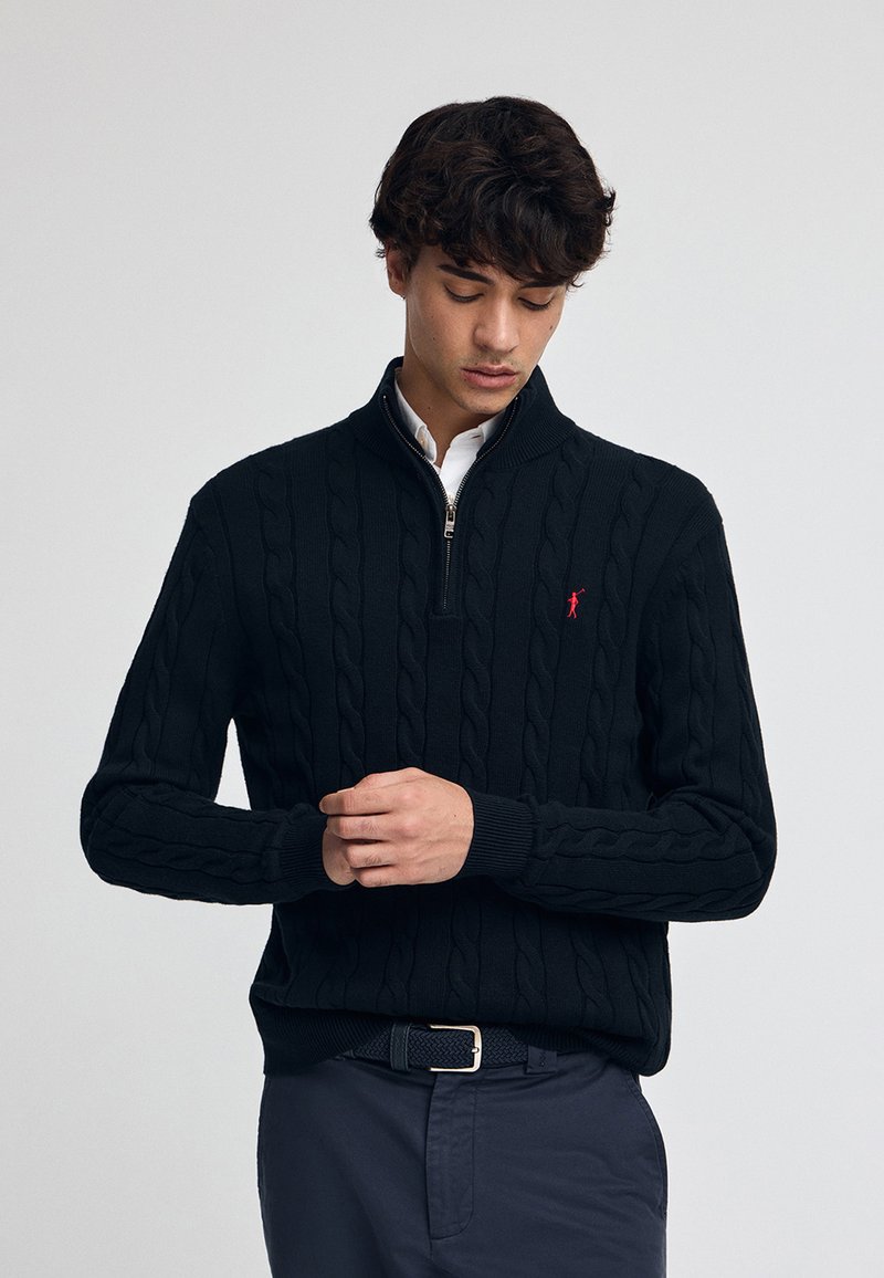 Pull noir en tricot torsadé avec un col zippé, arborant un petit logo rouge sur la poitrine et des poignets côtelés, associé à un pantalon foncé.