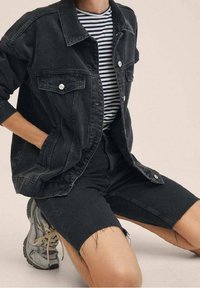 Persona inginocchiata che indossa una giacca di jeans nera, pantaloni corti neri sfrangiati, una maglietta a righe bianco e nera e scarpe da ginnastica grigie.