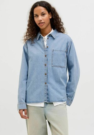 Camisa de mezclilla azul claro con botones al frente, mangas largas y un bolsillo en el pecho. Presenta botones marrones y un corte relajado.