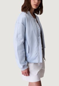 Jasnoniebieska kurtka bomber wykonana z gładkiej tkaniny, z zamkiem błyskawicznym z przodu, prążkowanymi mankietami i bocznymi kieszeniami. Luźny krój w minimalistycznym stylu.