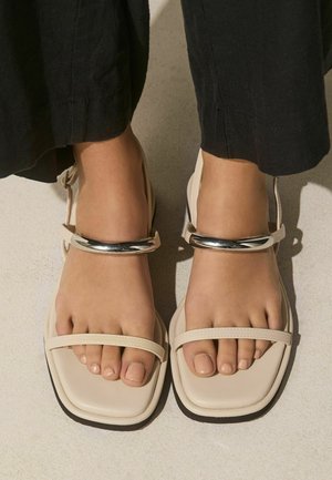 Feet wearing beige square-toe sandals with thin straps and metallic ankle accents, paired with black cropped pants.