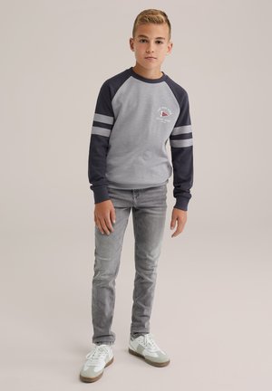 Sudadera gris de manga larga con mangas raglán azul marino, con rayas plateadas y un logo, combinada con jeans gris claro y zapatillas blancas.